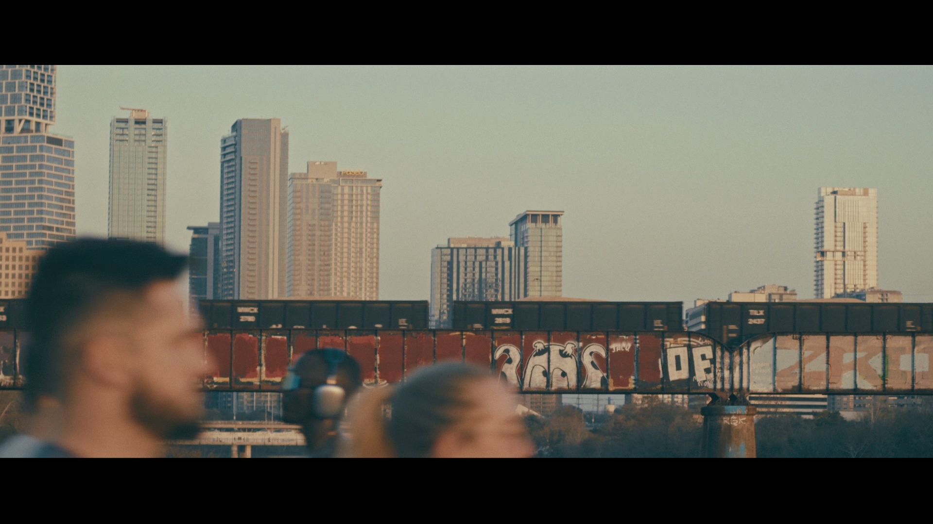 Blurred people in front of a city skyline with tall buildings and a graffiti-covered train bridge in the background.