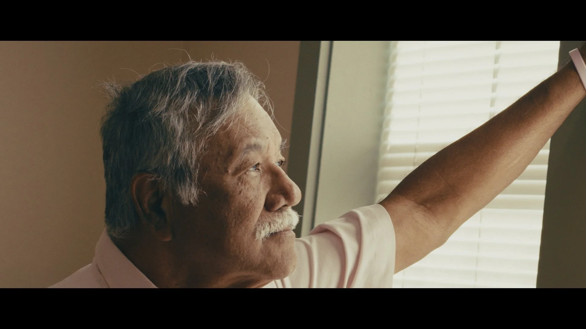An elderly man with gray hair and glasses reaches towards a window with sunlight coming through blinds.