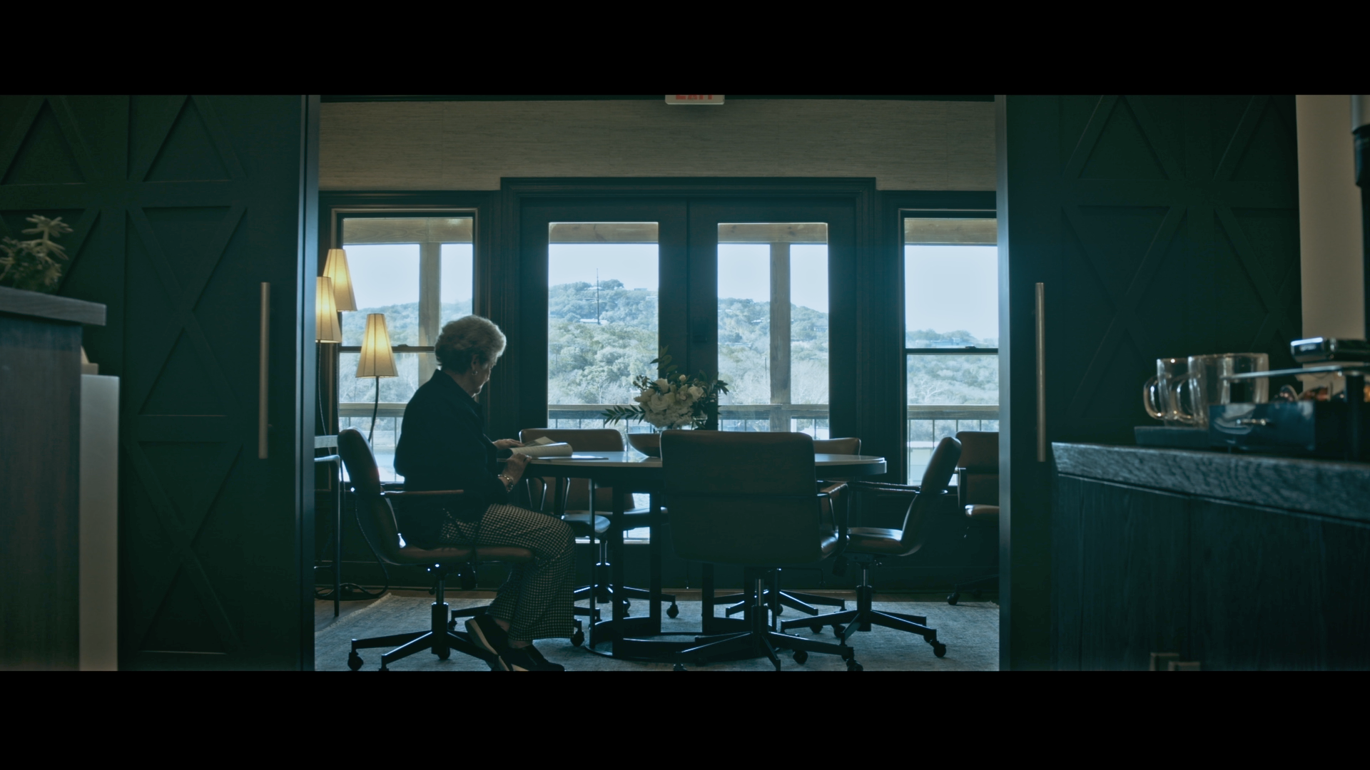 An older woman sits alone at a round dining table, reading a book or menu, with large windows showing a view of hills and trees in the background, in a bright room with modern decor.