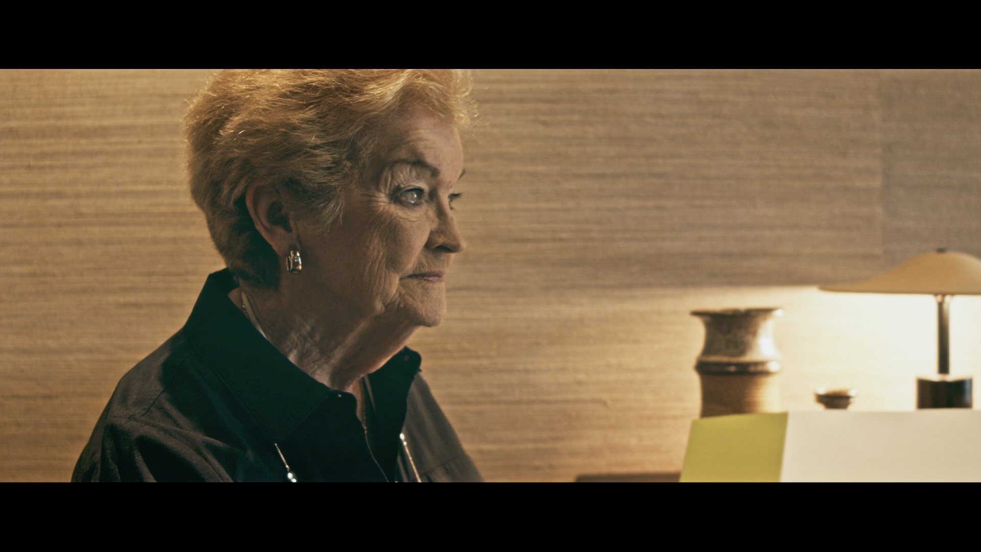 A close-up side profile of an elderly woman with short, curly, blonde hair wearing earrings, sitting at a table with a lamp and a yellow notebook, against a textured beige wall.