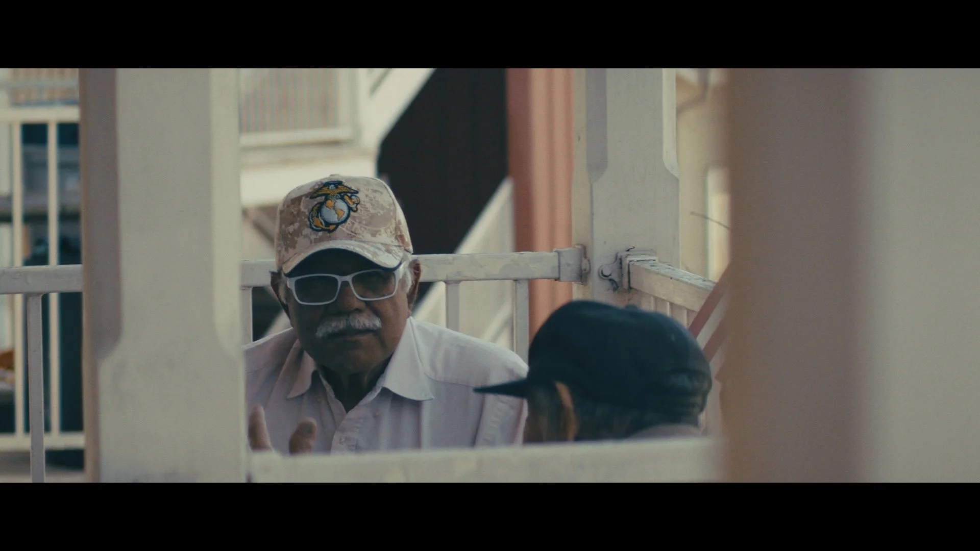 An elderly man with a gray mustache and sunglasses wearing a cap with a military insignia, engaged in conversation with a person wearing a black cap outside next to a white fence.