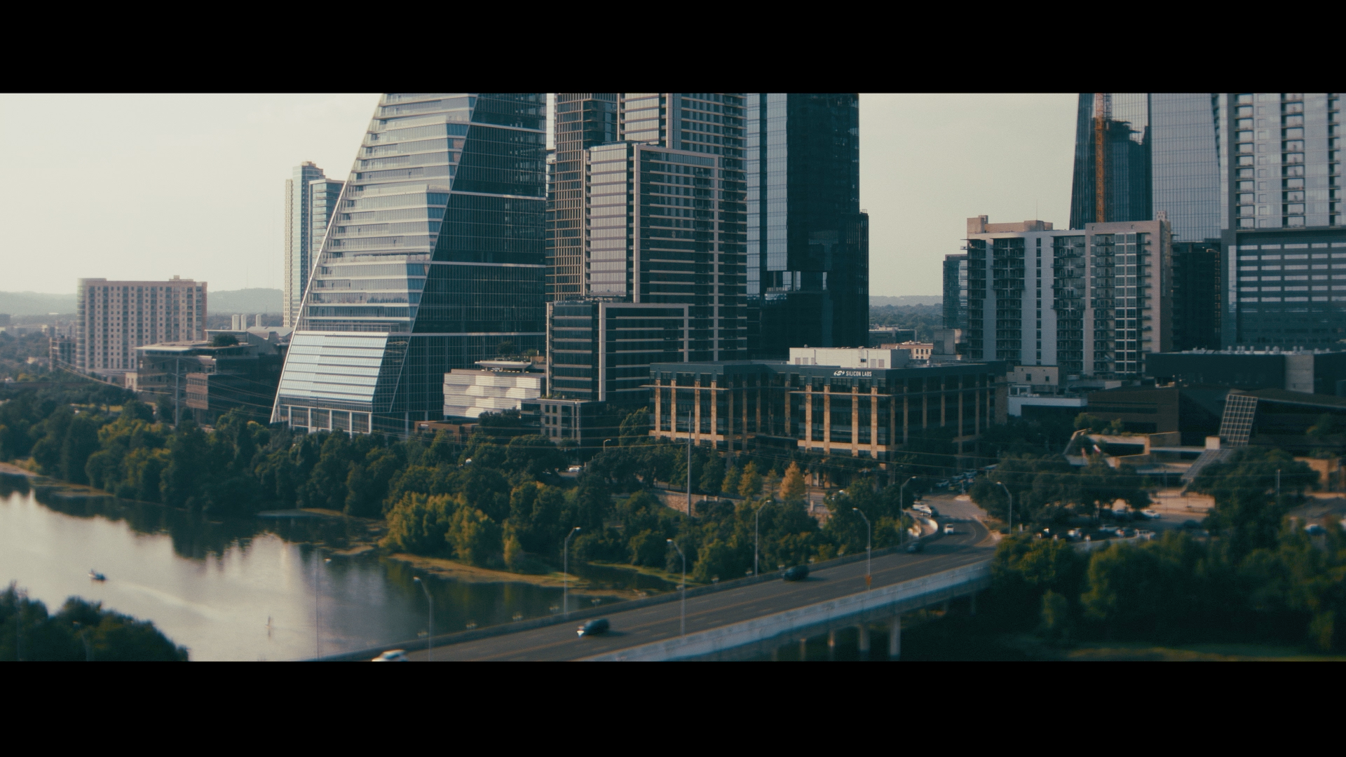 City skyline with modern skyscrapers along a river, trees, and a highway in the foreground.