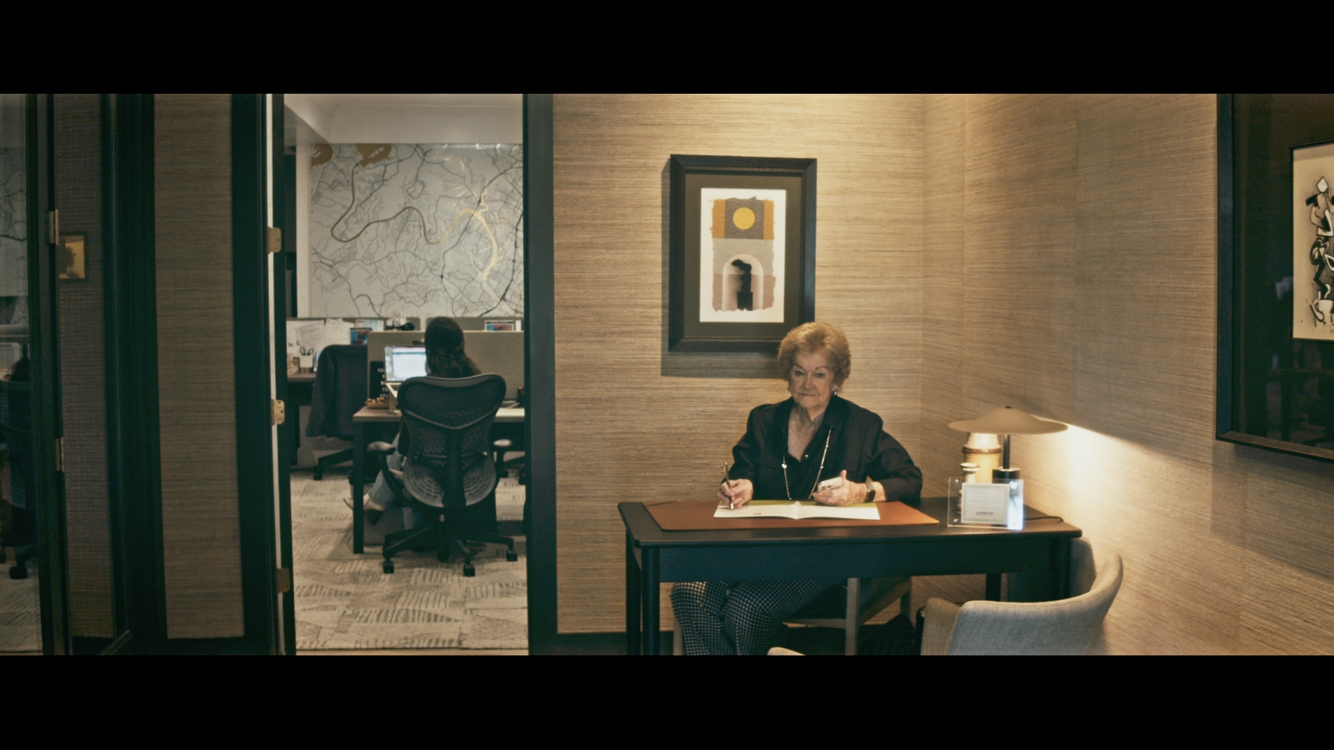 Office space with an older woman sitting at a desk, writing in a notebook, with a lamp and framed artwork on the wall. In the background, a person is working at a computer in a separate room.