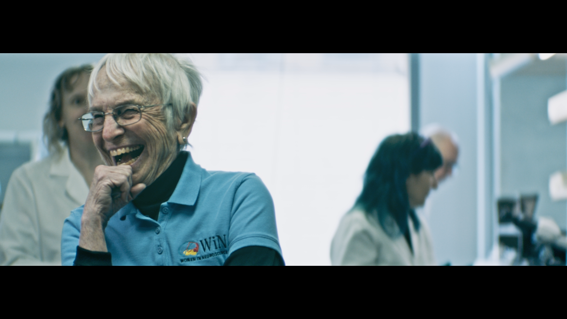 Women in neuroscience conference sharing a laugh, with focus on an older woman with short blonde hair and glasses.
