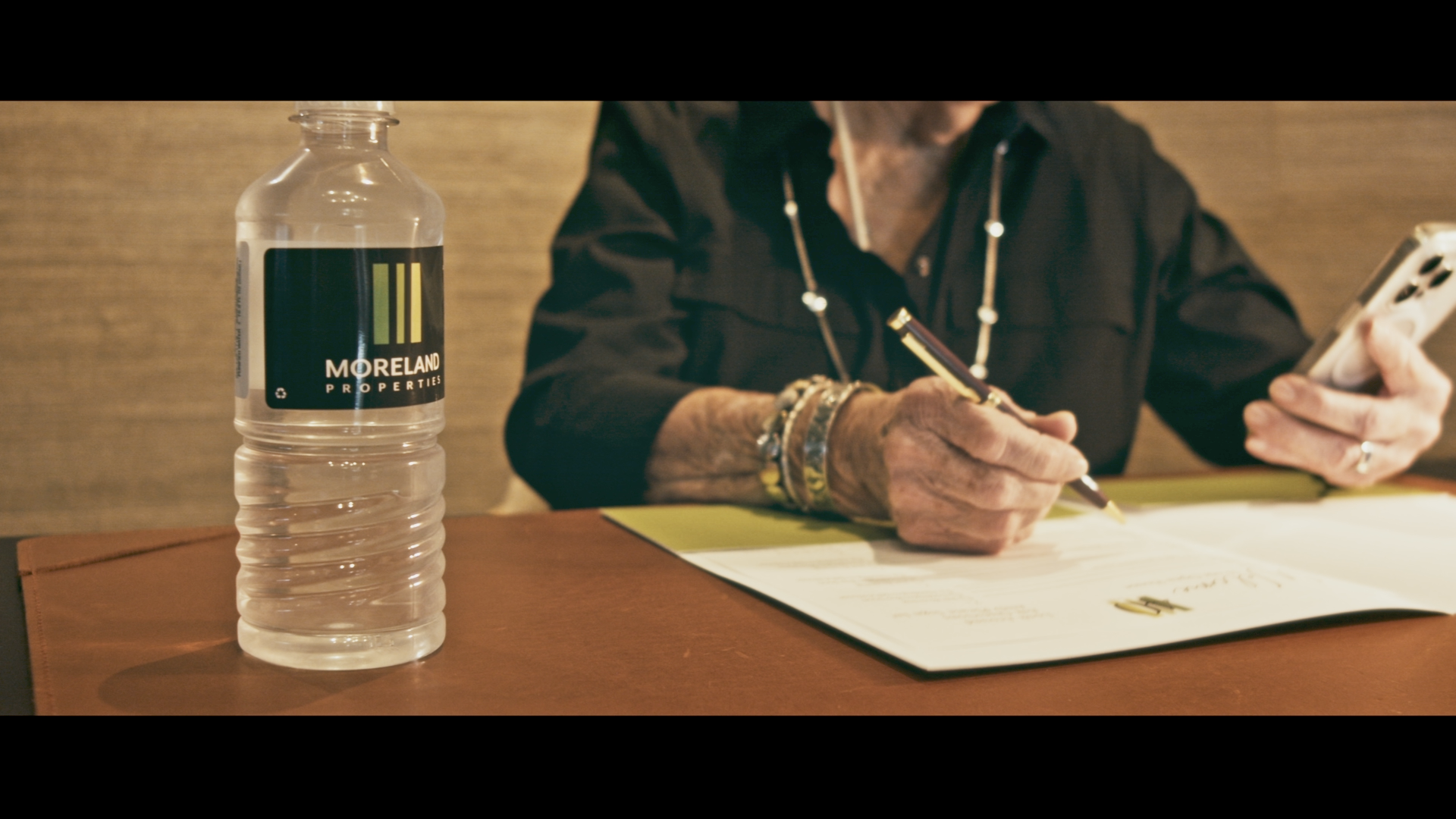 Close-up of a water bottle labeled 'Moreland Properties' on a table. In the background, a person wearing jewelry is writing on a document and looking at their phone.