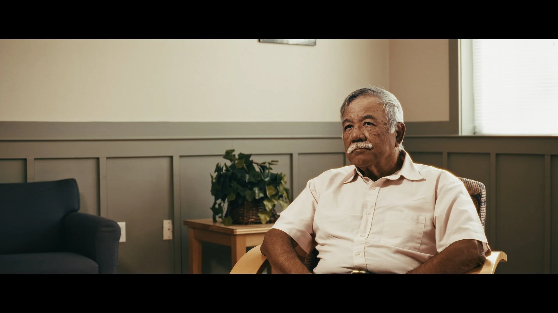 A senior man with gray hair and a mustache sitting in a waiting room or office, looking thoughtfully to the side. He is wearing a light-colored short-sleeve shirt, with a potted plant on a table behind him. The room has beige walls, a window with blinds, and some electrical outlets visible.