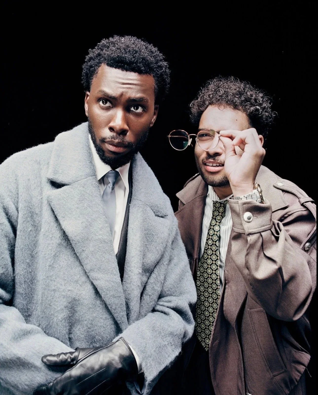 Two men with curly hair and glasses, dressed in vintage-style clothing, standing against a black background. One man is wearing a gray coat and a white shirt, the other a beige trench coat and a patterned tie, both looking serious or curious.