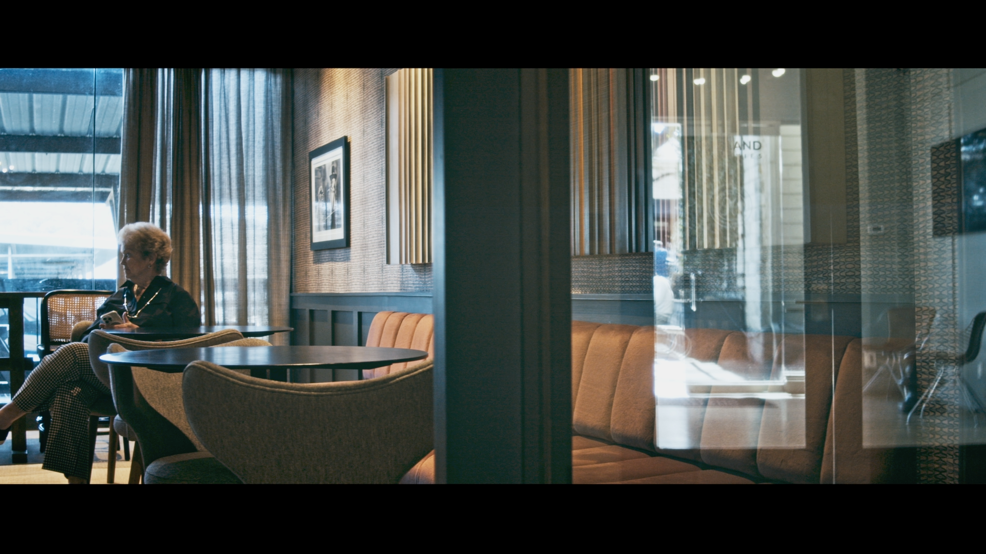 Interior of a modern coffee shop or lounge with a woman sitting on a chair, looking at her phone. The space features warm lighting, abstract art on the walls, large windows with curtains, and contemporary furniture including cushioned chairs and tables.