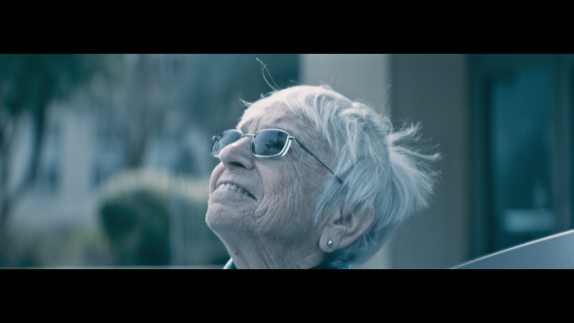 Close-up of an elderly woman with gray hair, wearing sunglasses, smiling and looking up outdoors.