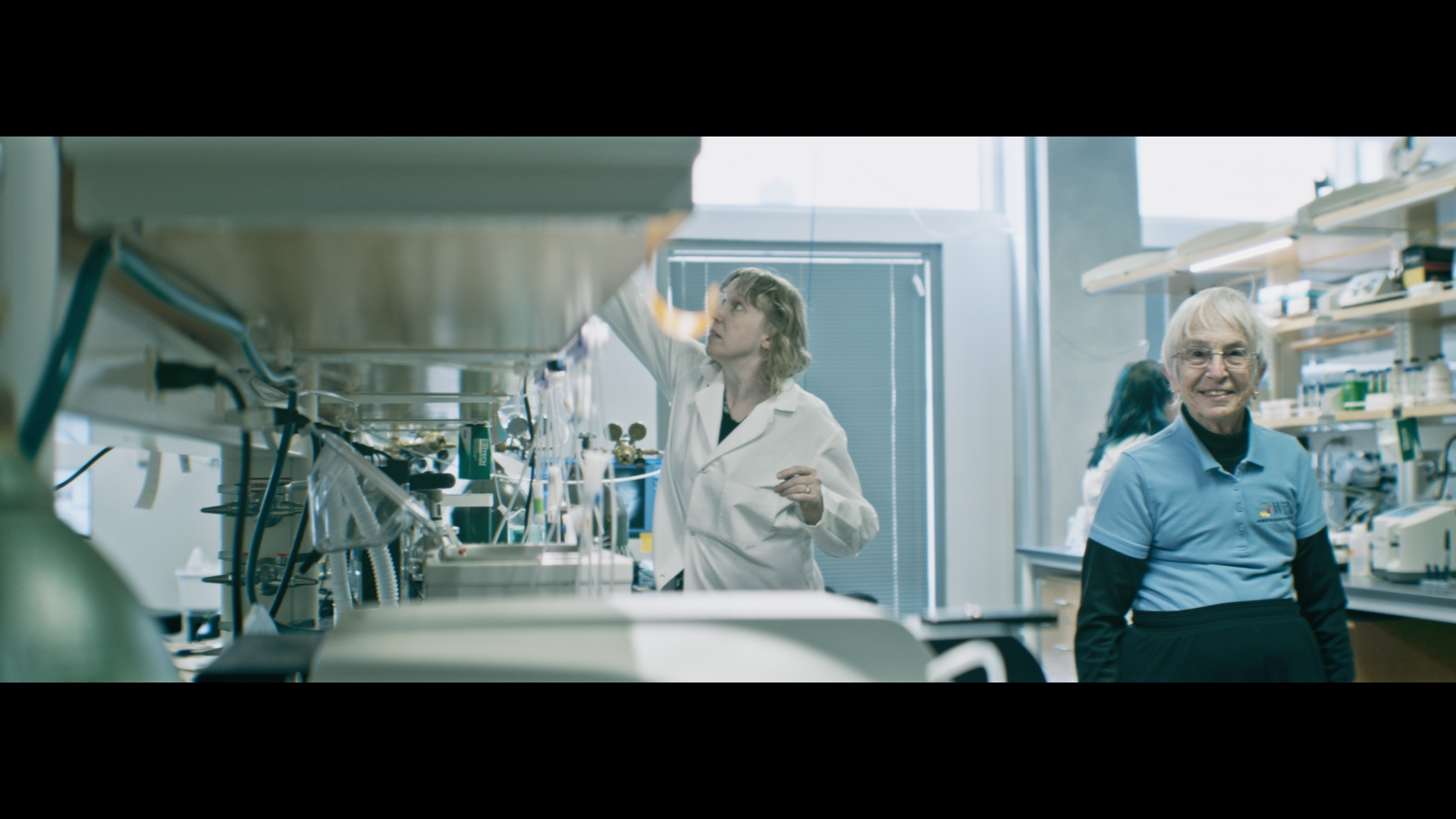 Scientists working in a laboratory, with one woman in a white lab coat handling equipment, and an older woman smiling in a blue shirt in the background.