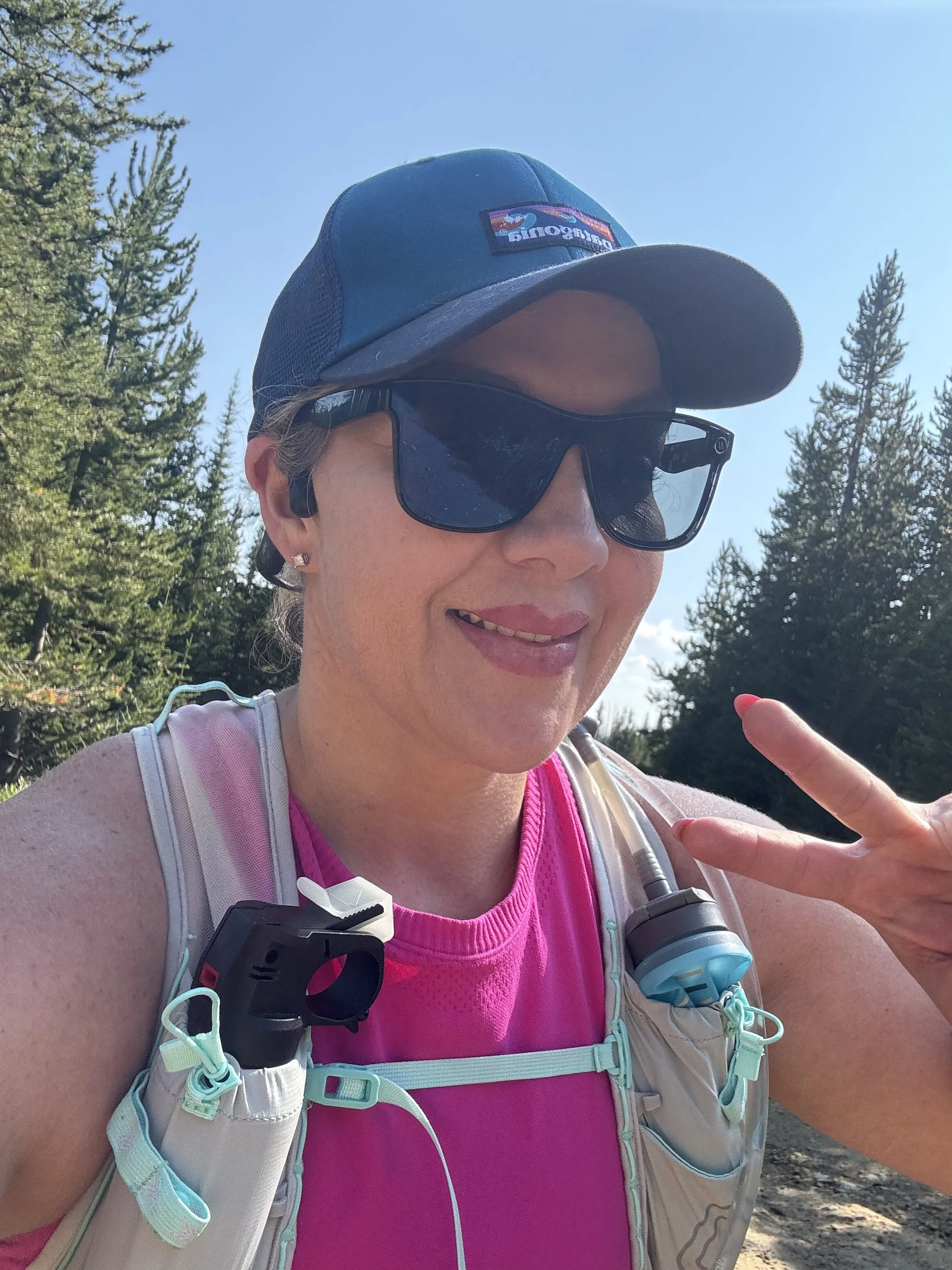 Woman in pink running gear outdoors making a peace sign, wearing sunglasses and a cap, with trees and blue sky in the background.