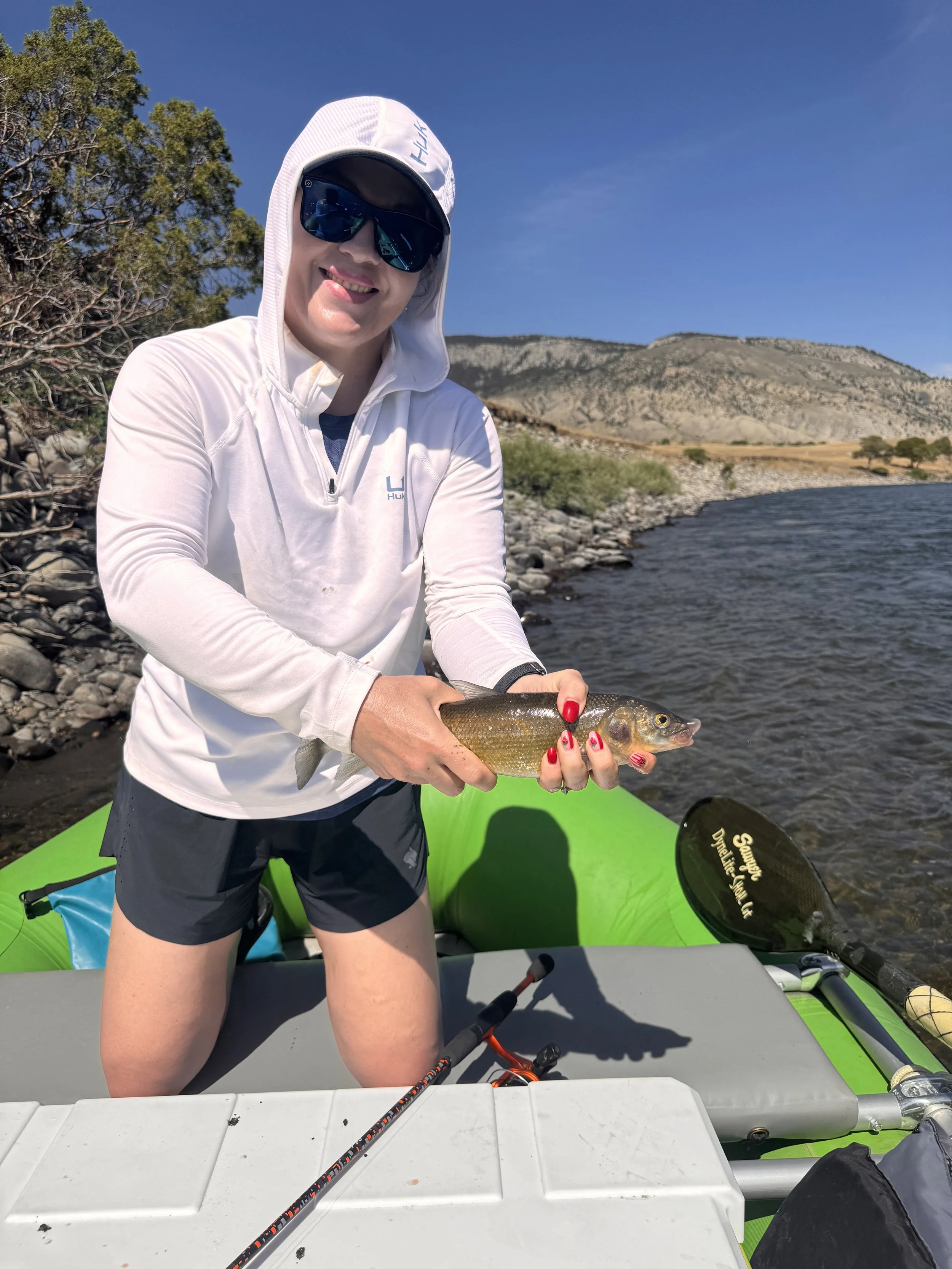 Woman in white hoodie and sunglasses holding a fish on a kayak by a river with mountains in the background