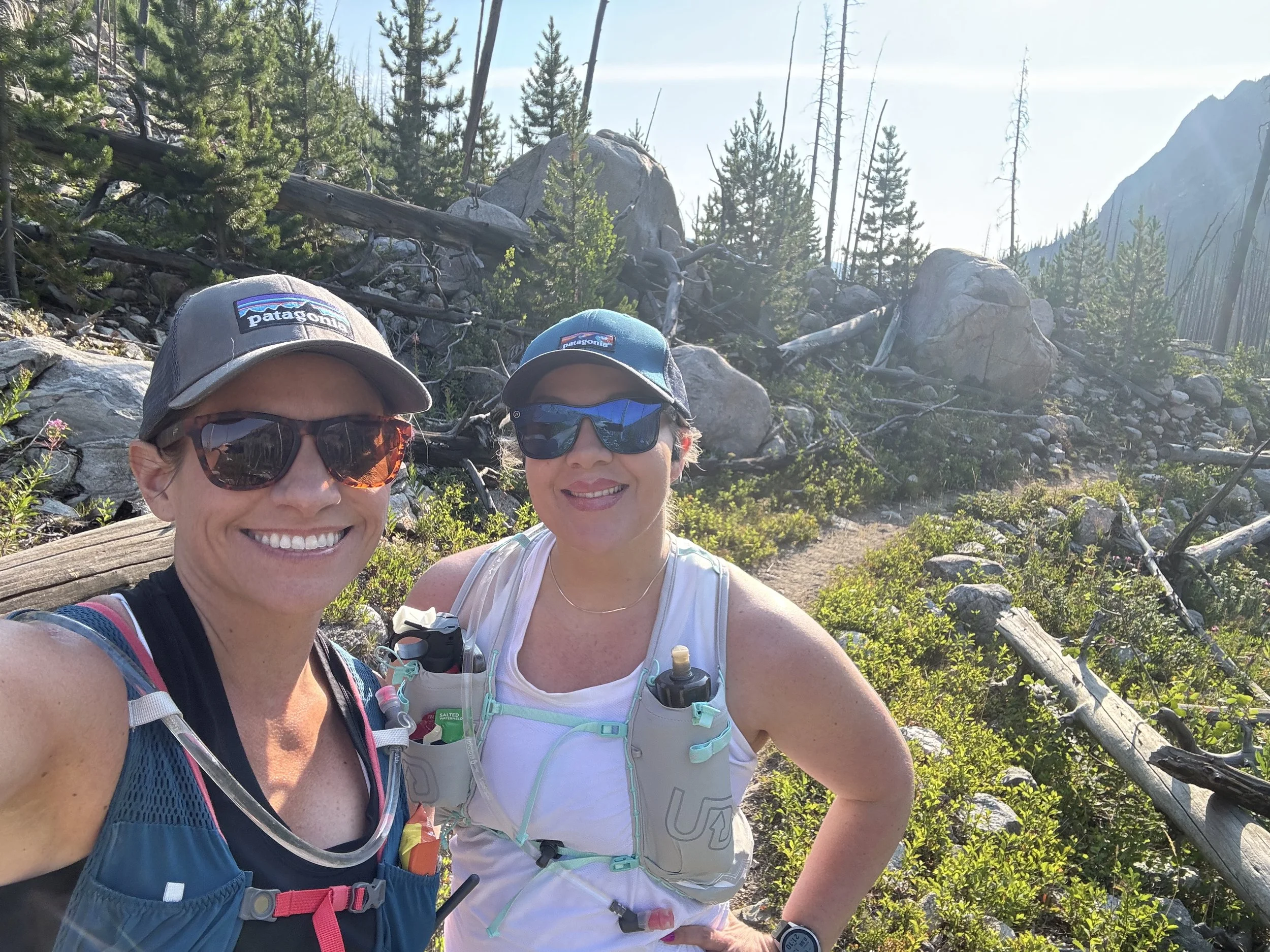 Two women wearing sunglasses and hats taking a selfie outdoors on a trail surrounded by green trees, rocks, and fallen logs in a mountainous area.