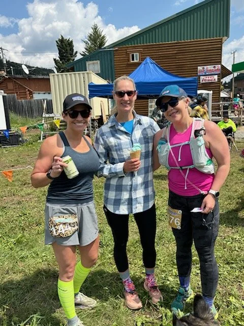 Three women standing outdoors at a running event, holding ice cream cones and drinks, dressed in athletic gear and sunglasses, with tents and other participants in the background.