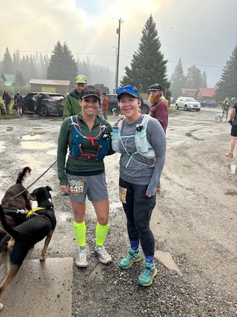Two smiling women in running gear and hydration packs standing on a wet, gravel trail in a forested mountain area with parked cars and other runners in the background, accompanied by a dog on a leash.