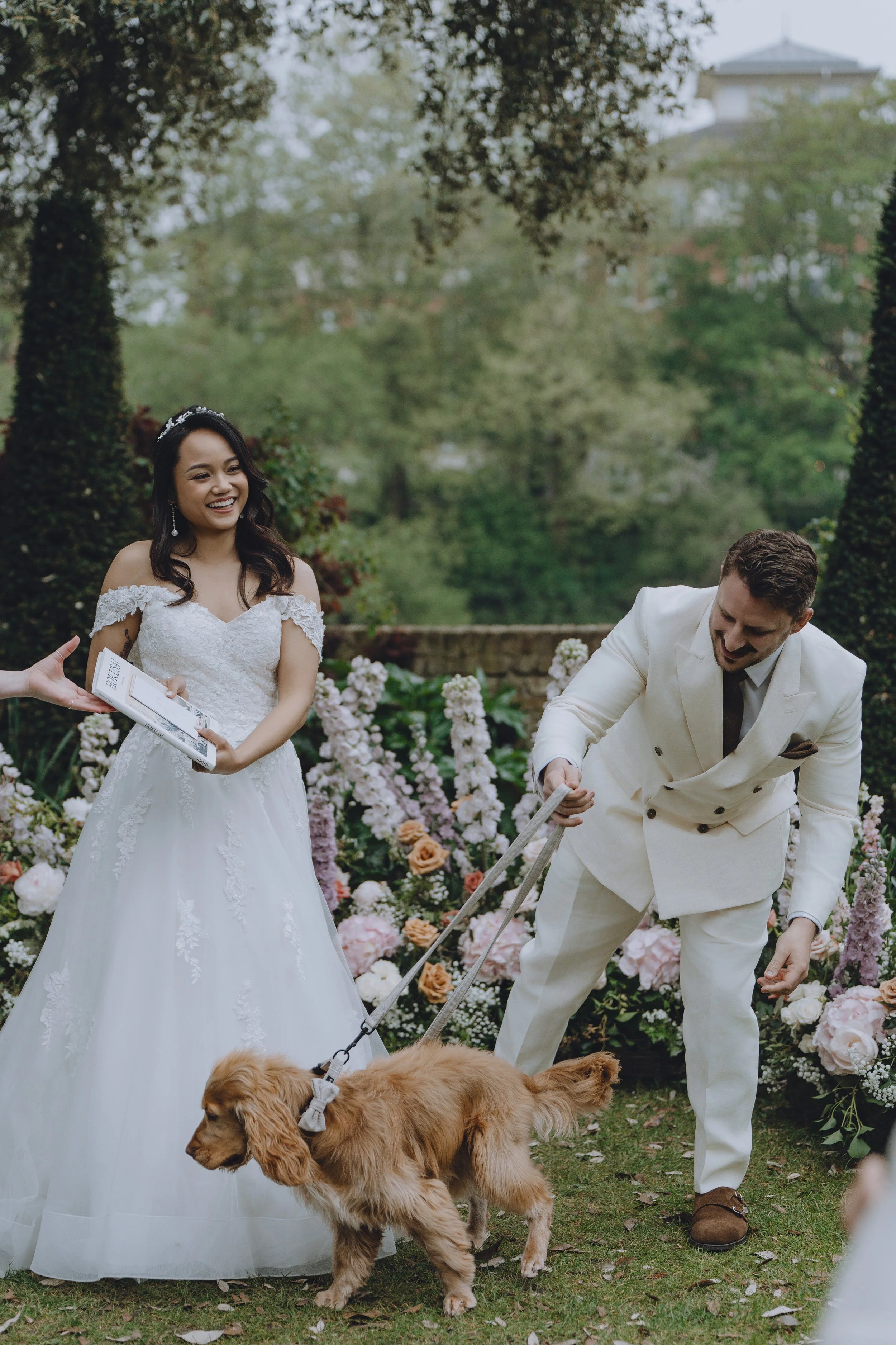 A bride in a white wedding gown and a tiara smiling while holding a small object, possibly a book or card, standing outdoors among colorful flowers with a groom in a cream suit and black tie, who is bending down near a golden retriever dog.