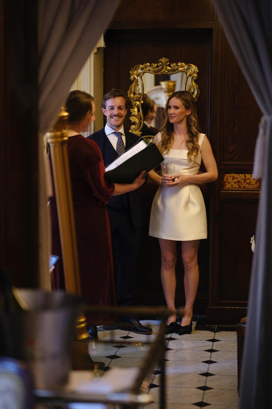 A woman and man dressed formally, standing in front of a woman holding a script, in an ornate room with wood paneling and a gilded mirror, at a wedding or ceremony.