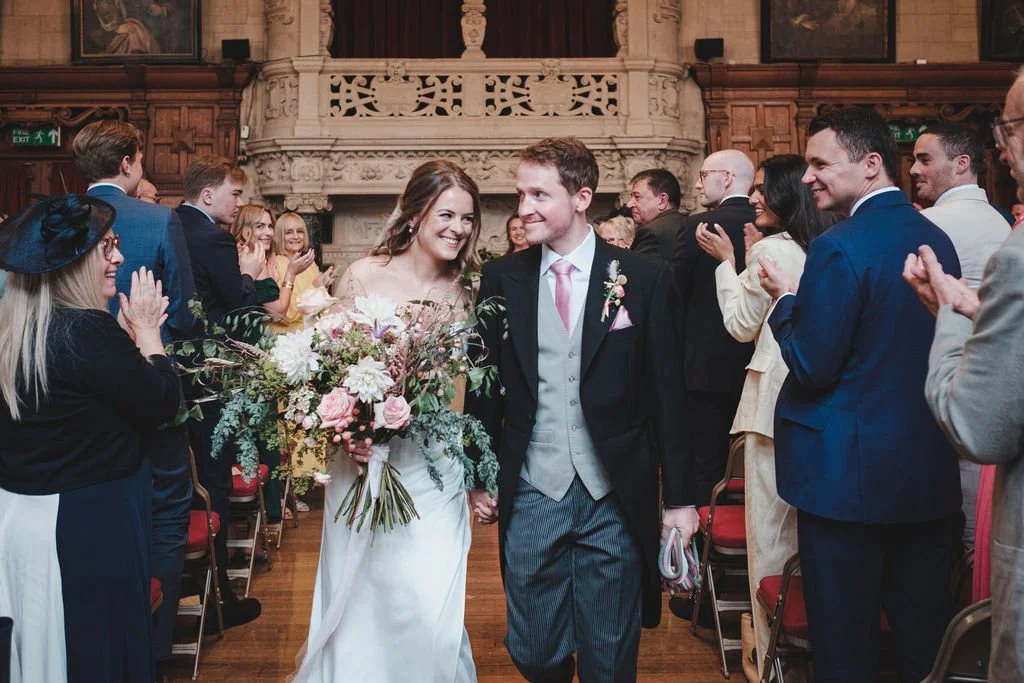 A bride and groom walking down the aisle, smiling, surrounded by clapping guests in a decorated indoor venue.