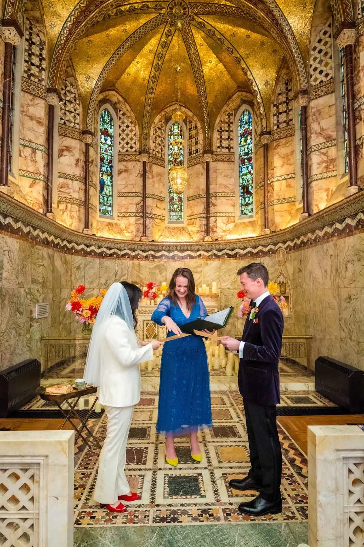 A wedding ceremony taking place inside a richly decorated church with stained glass windows and an ornately gold-leafed vaulted ceiling. The bride and groom are facing each other, with an officiant in a blue dress standing between them. The bride wears a white suit with a white veil and red shoes. The groom wears a dark suit with a boutonniere. There are flowers and candles in the background.