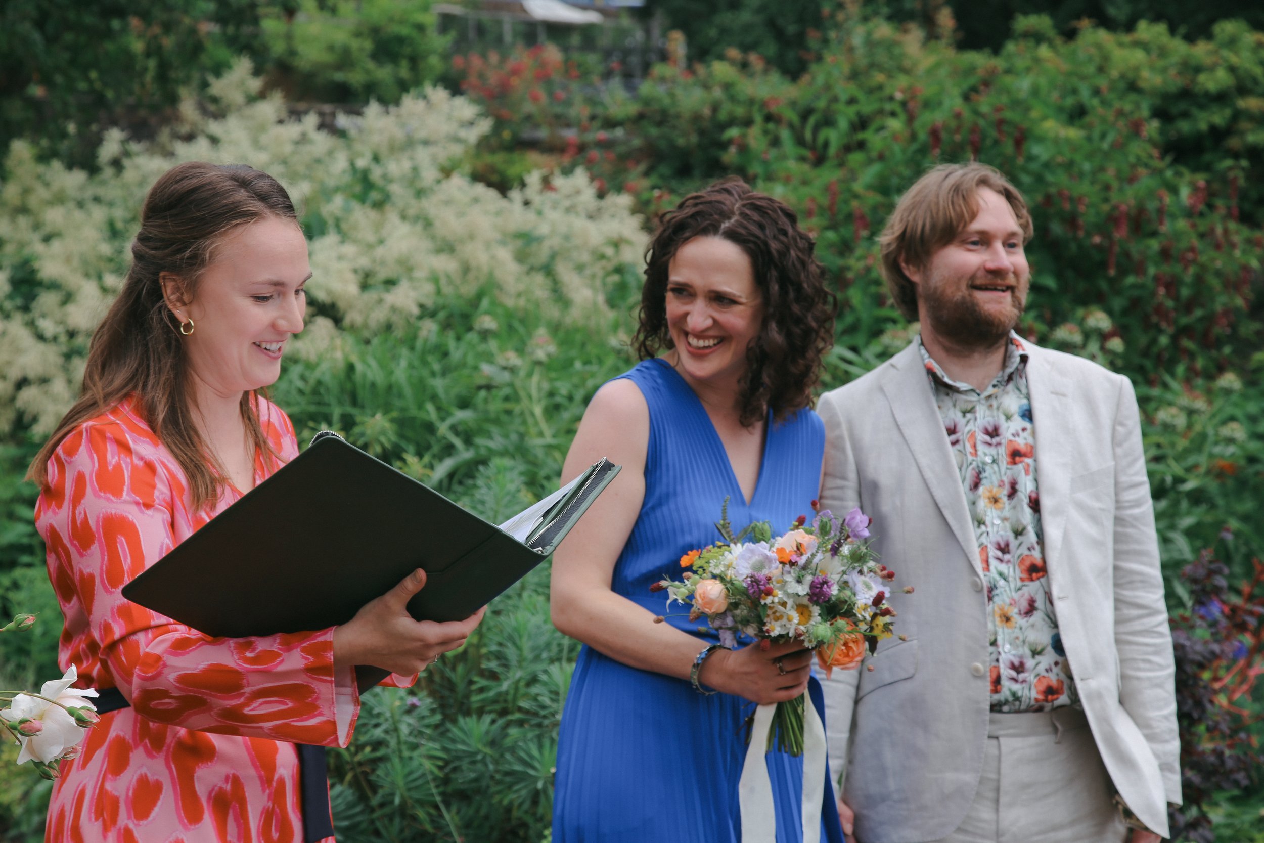 Three people at a garden wedding ceremony. The woman on the left is reading from a black folder, the woman in the middle is holding a bouquet of flowers, and the man on the right is smiling. All are dressed in colorful attire.