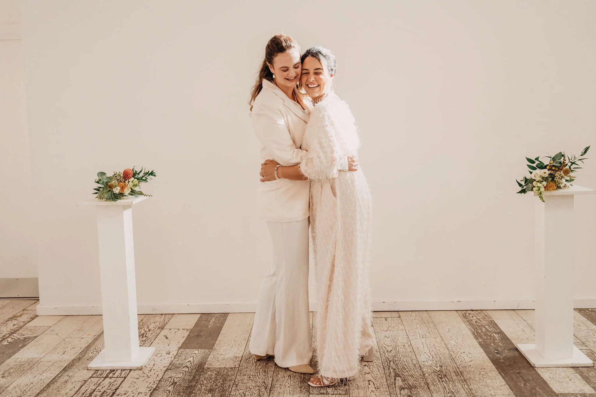 Two women, one dressed in white and the other in beige, are embracing each other and smiling in a minimalist indoor setting with floral arrangements on white pedestals.