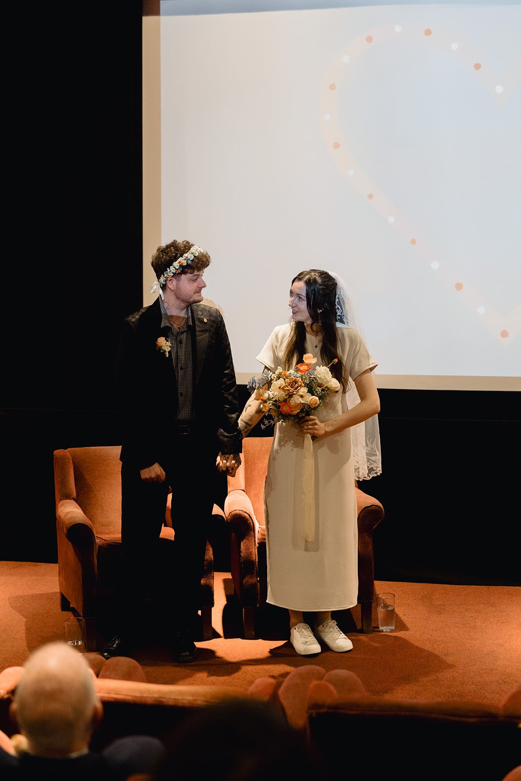 A couple dressed in wedding attire holding hands on stage, with the woman holding a bouquet of flowers and wearing a veil, and a man with a flower crown, standing in front of a large screen. Audience members are visible in the foreground.
