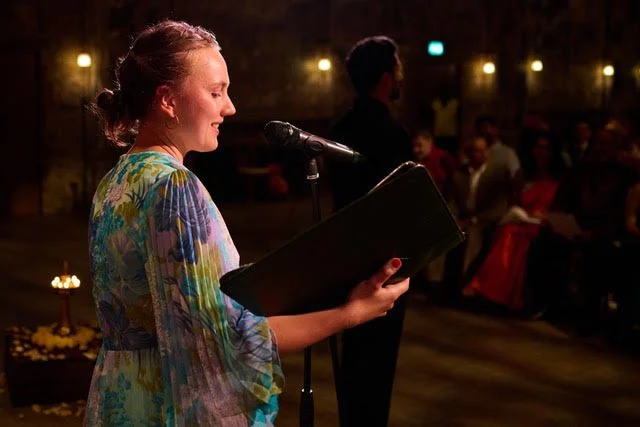 A woman with red hair reading from a black book at a microphone during a performance or event, with an audience seated in the background.