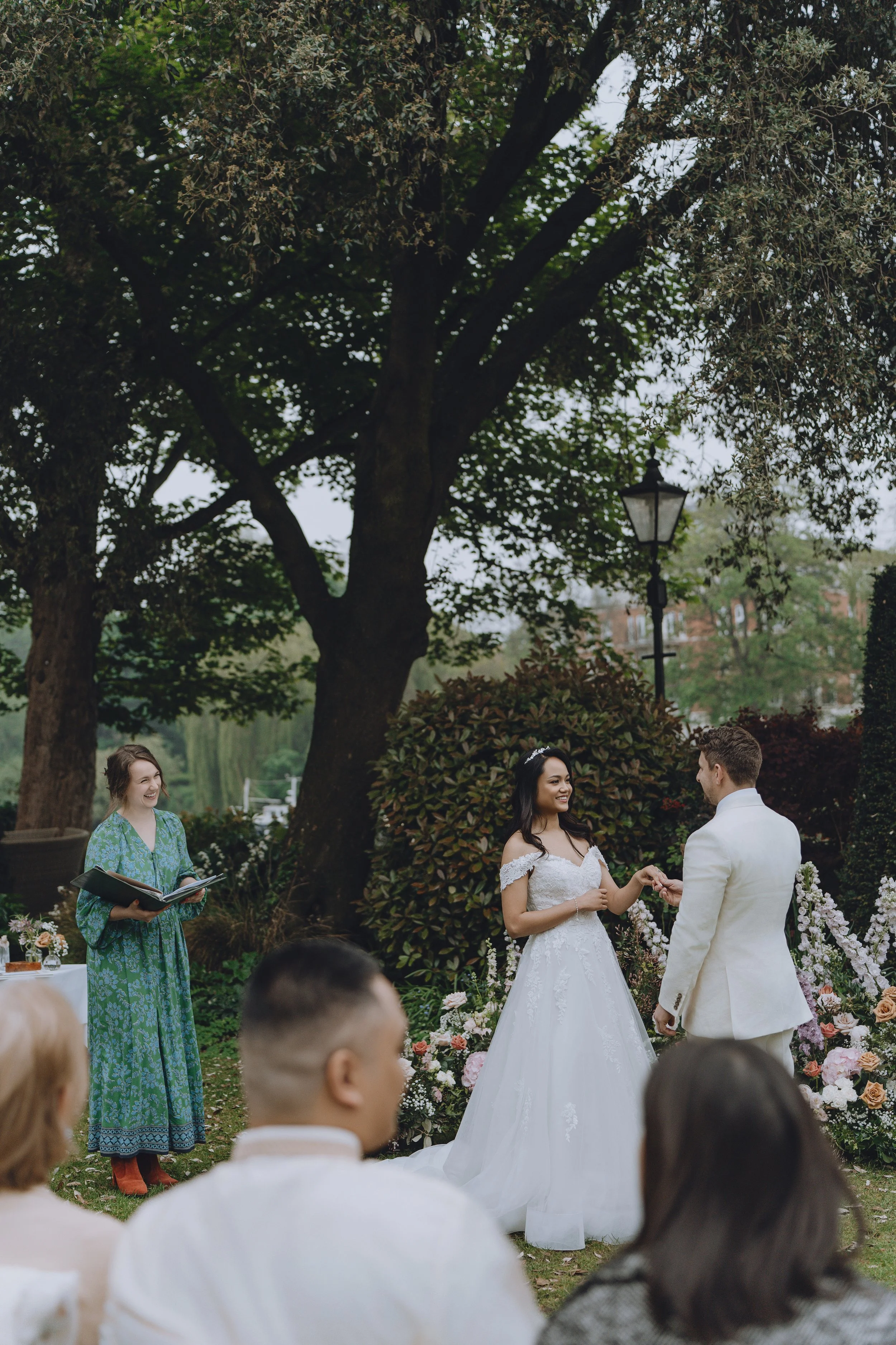 A wedding ceremony taking place outdoors under large trees, with a bride and groom exchanging vows, surrounded by floral arrangements and guests.