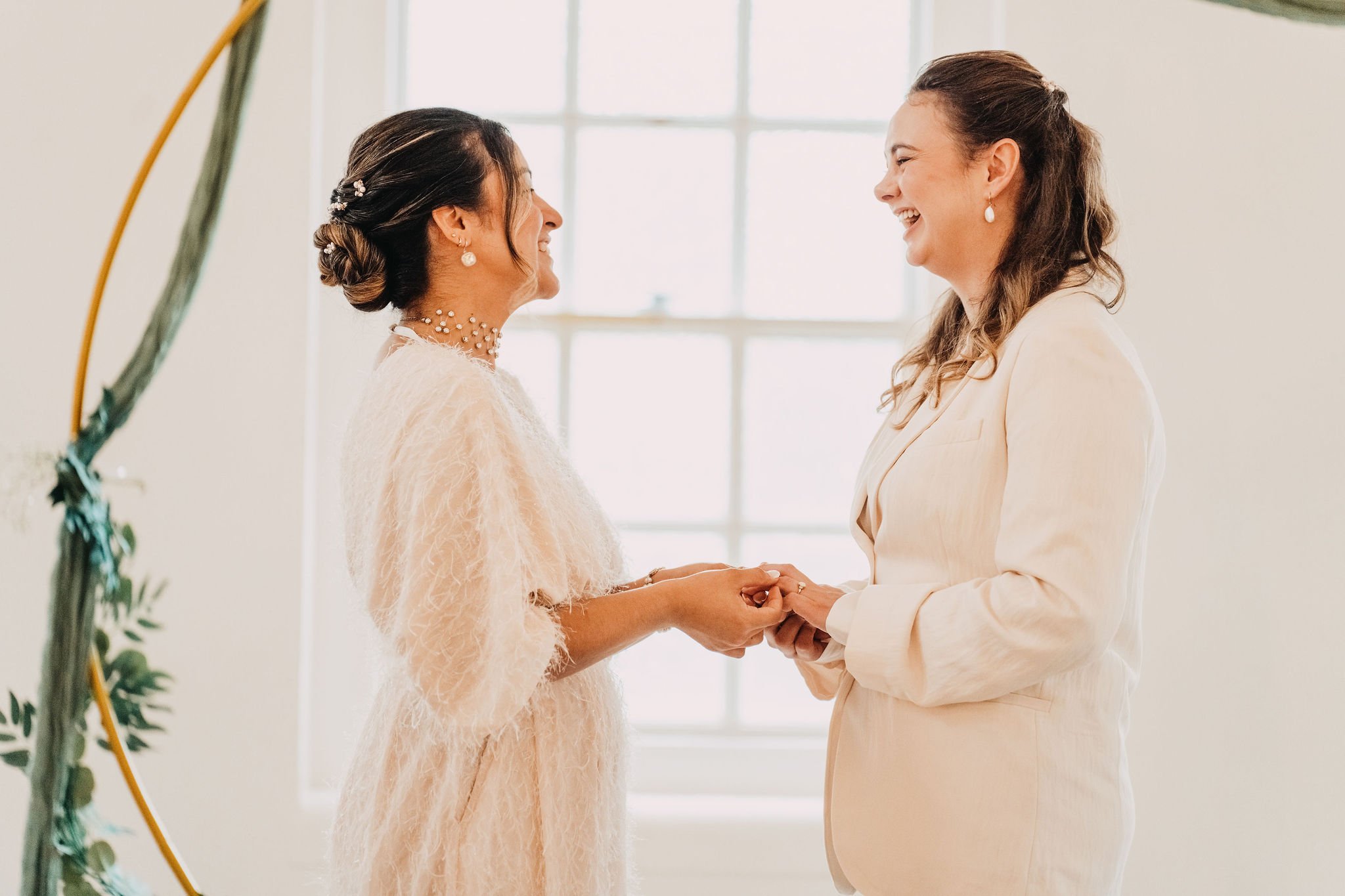 Two women dressed in elegant clothing holding hands and facing each other with smiling expressions in front of a window.