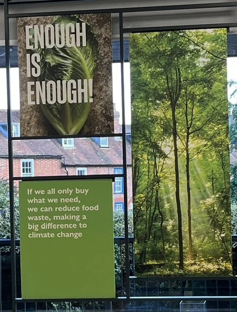 Three green signs hanging in supermarket window, one with trees, one with Enough is Enough and the third with If we all only buy what we need, we can reduce food waste, making a big difference to climate change