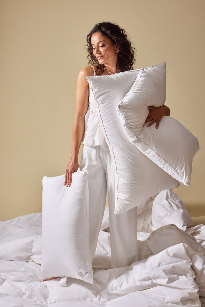 A smiling woman in white silk pajamas holding fluffy white down feathers pillows in bed, by Bakkes Images.