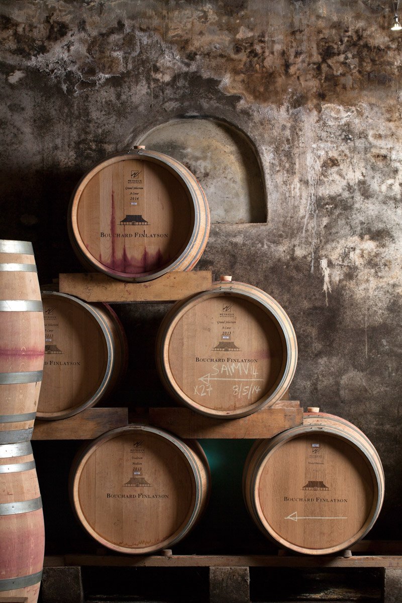 Stacked French oak wine aging barrels resting against a rustic stone cellar wall, shot by Bakkes Images.