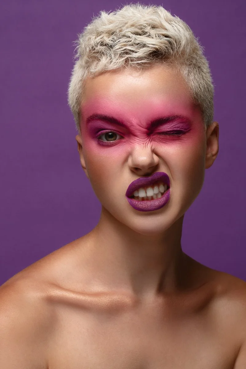 Playful high-fashion portrait of a model winking with intense pink eye and face makeup styling, by Bakkes Images.