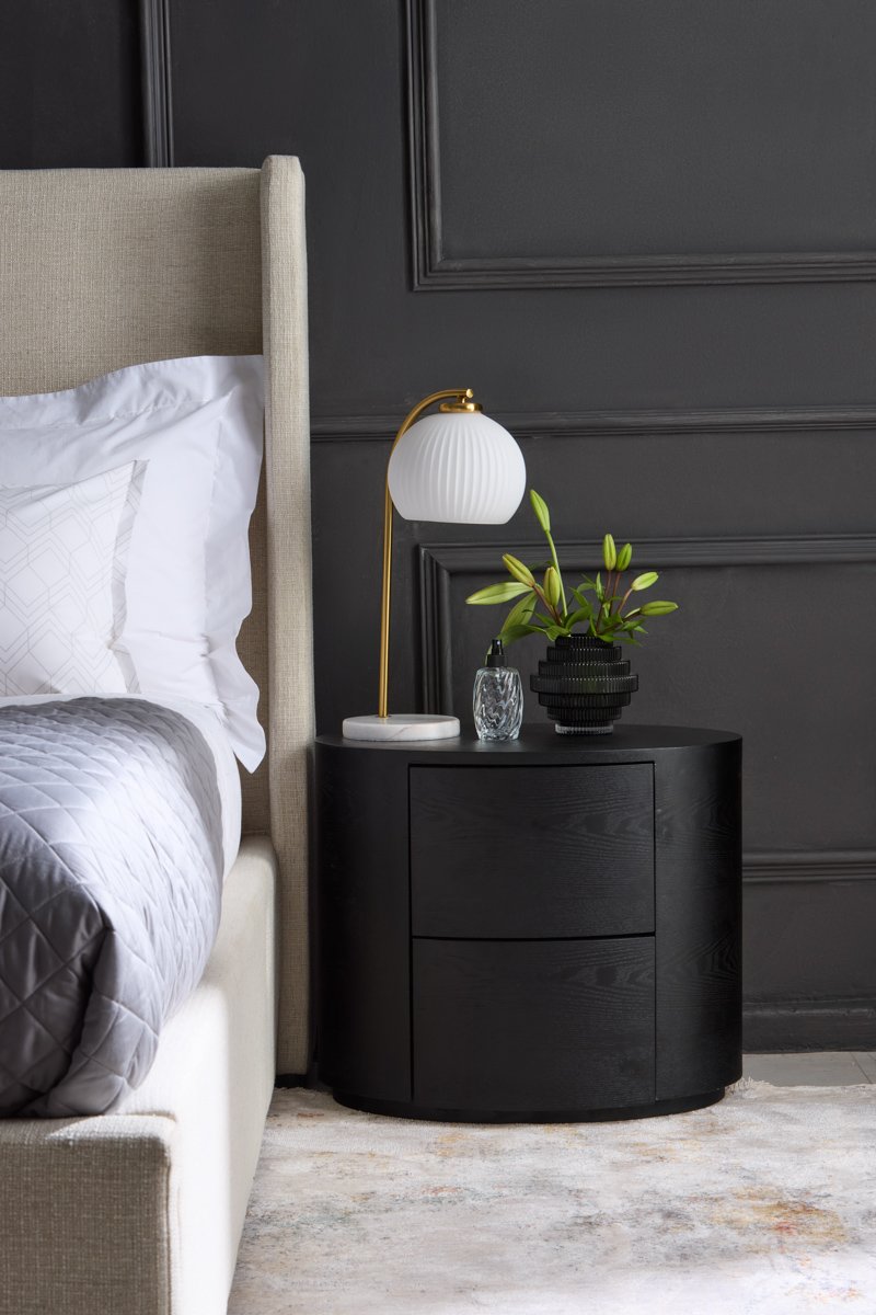 A round fluted black bedside table styled with a modern white dome lamp against grey walls, by Bakkes Images.