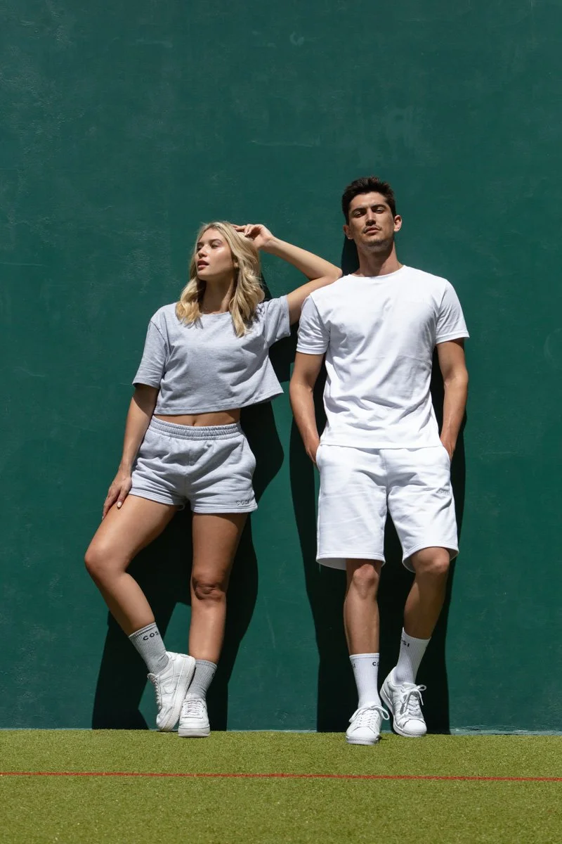Male and female models standing together against a solid green tennis court wall in Cosi apparel, by Bakkes.