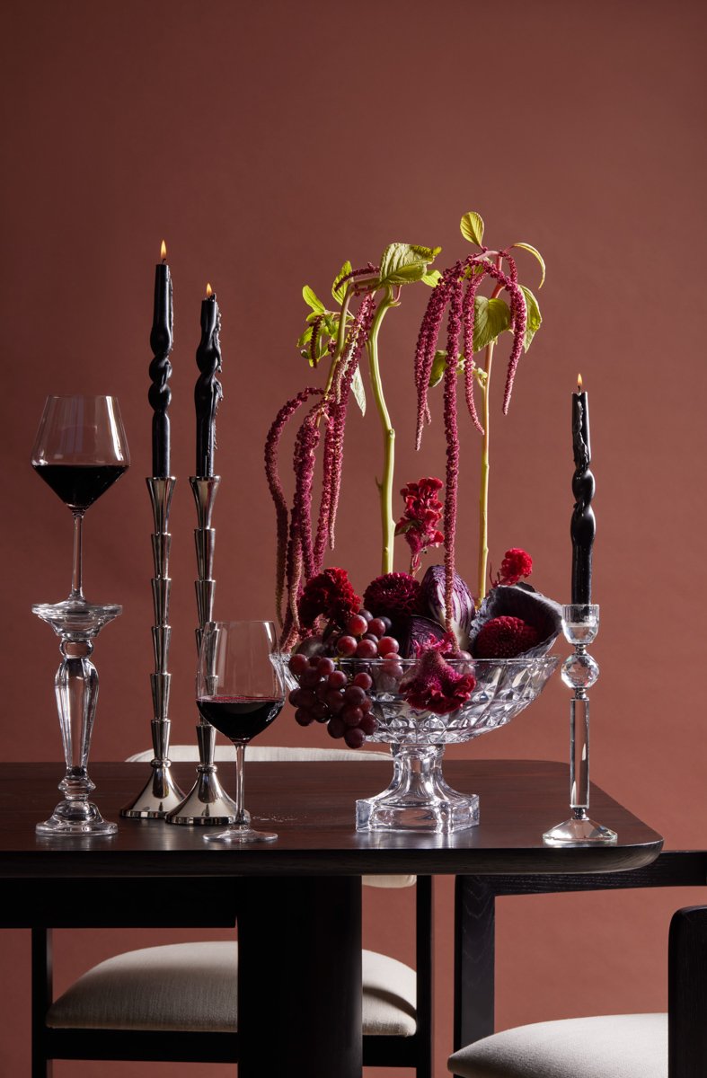 A dark dining room setup with an intricate floral centerpiece arrangement and tall black candles, by Bakkes.