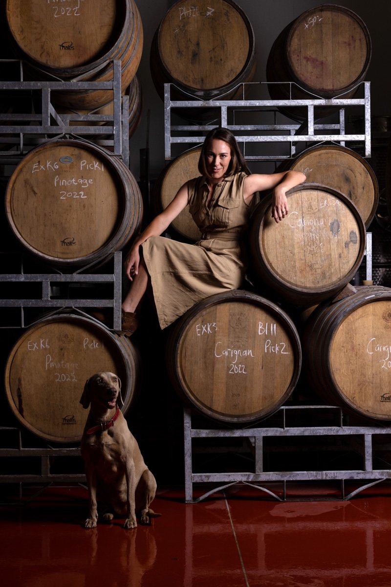 A woman sitting on a high stack of traditional oak barrels inside a moody maturation cellar, by Bakkes Images.