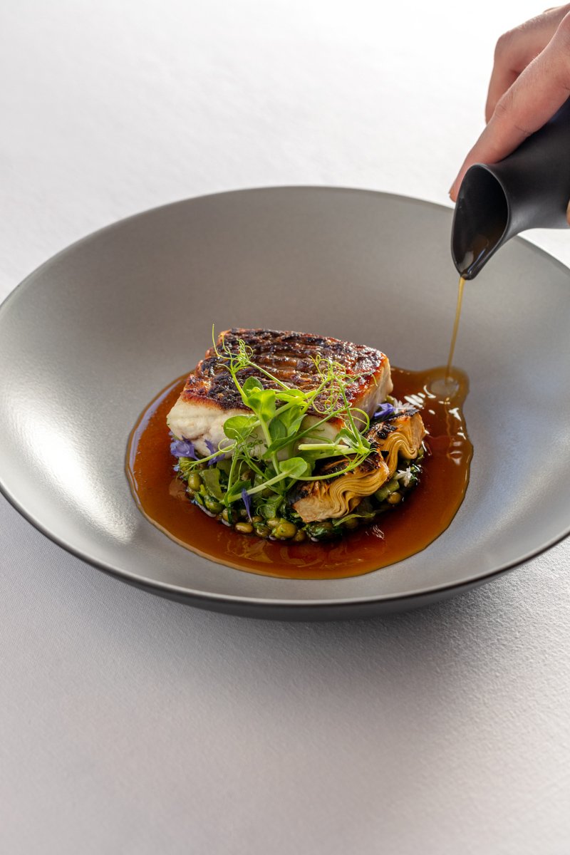 Action shot of rich jus being poured from a black jug over a seared fish dish in a modern grey bowl, by Bakkes.