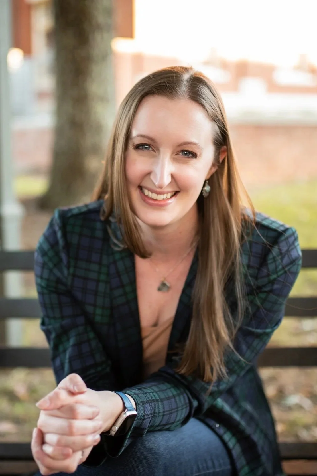 A smiling woman with long brown hair, wearing a plaid shirt and jewelry, sitting on a bench outdoors with trees and buildings in the background.