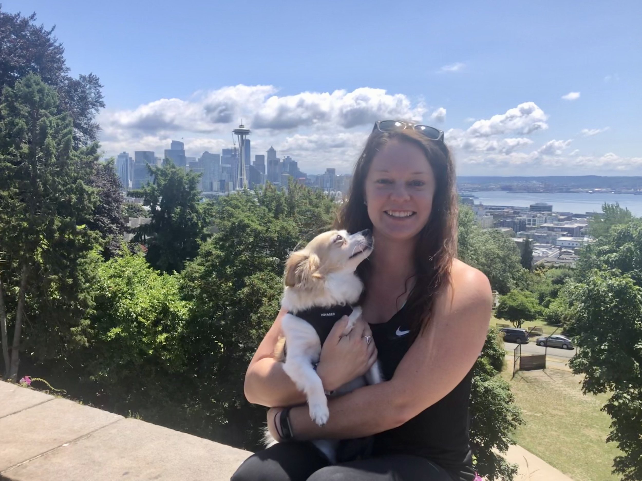 Woman holding a small dog in front of Seattle skyline with Space Needle, trees, and water in the background.