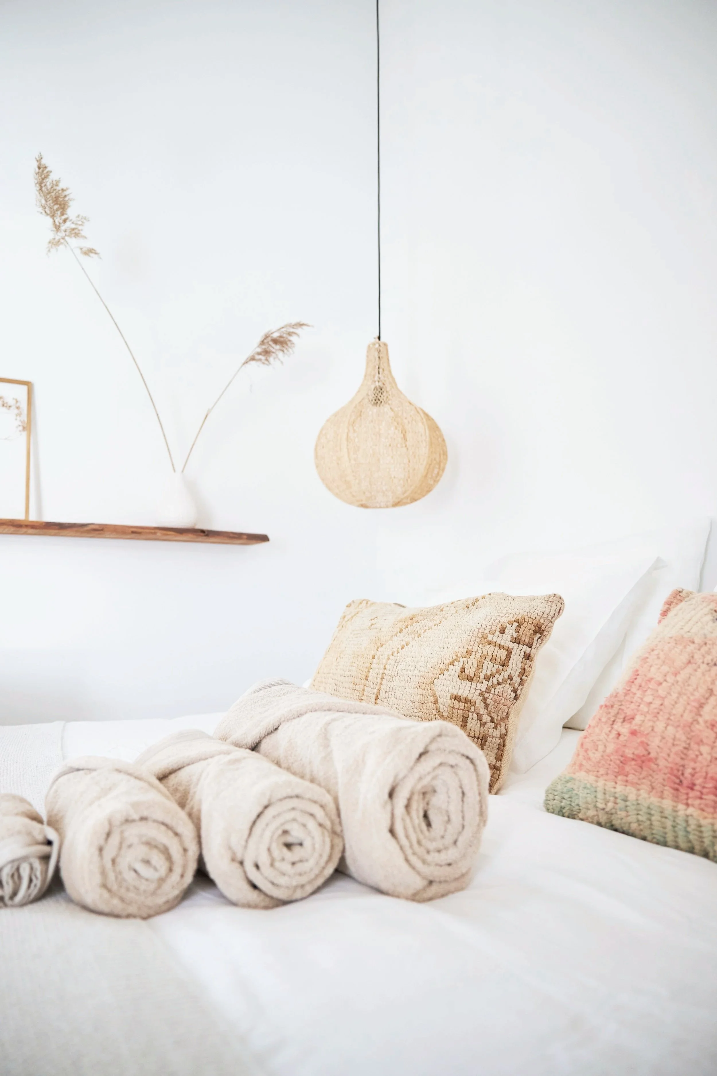 Decorative bedroom scene with rolled towels, pillows, wall shelf with vase and decorative pampas grass, hanging woven light fixture, and white wall.