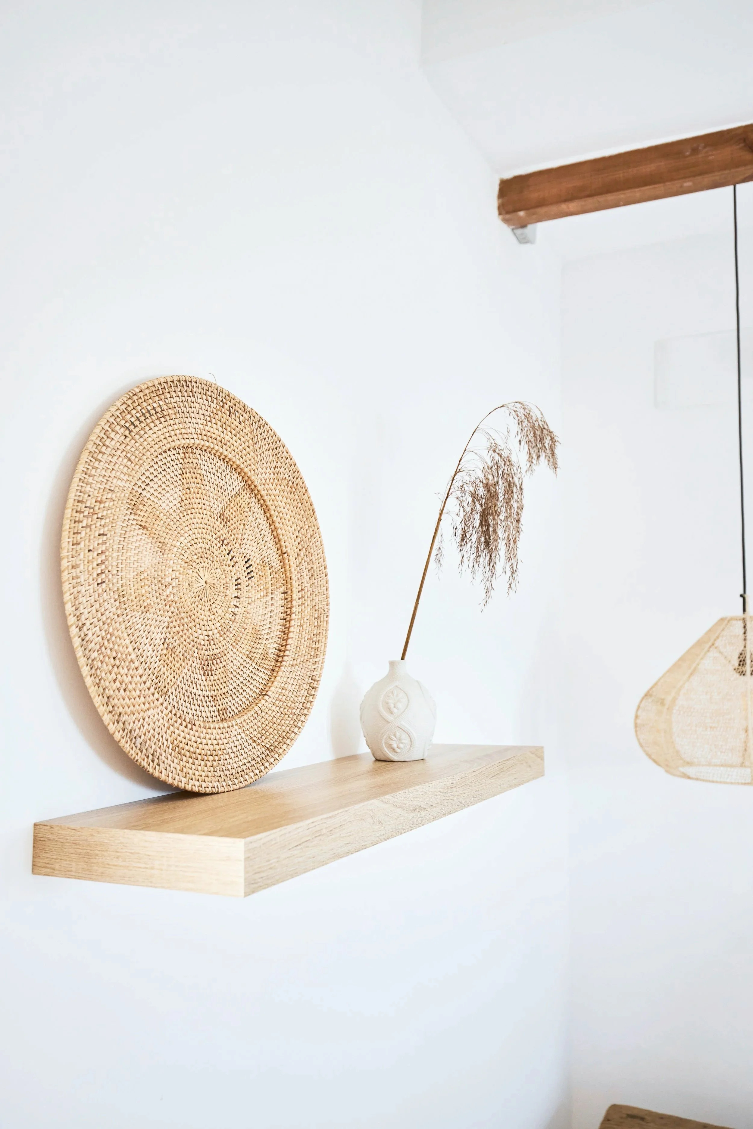 Decorative wicker plate and a white vase with dried grass on a wooden shelf against a white wall.
