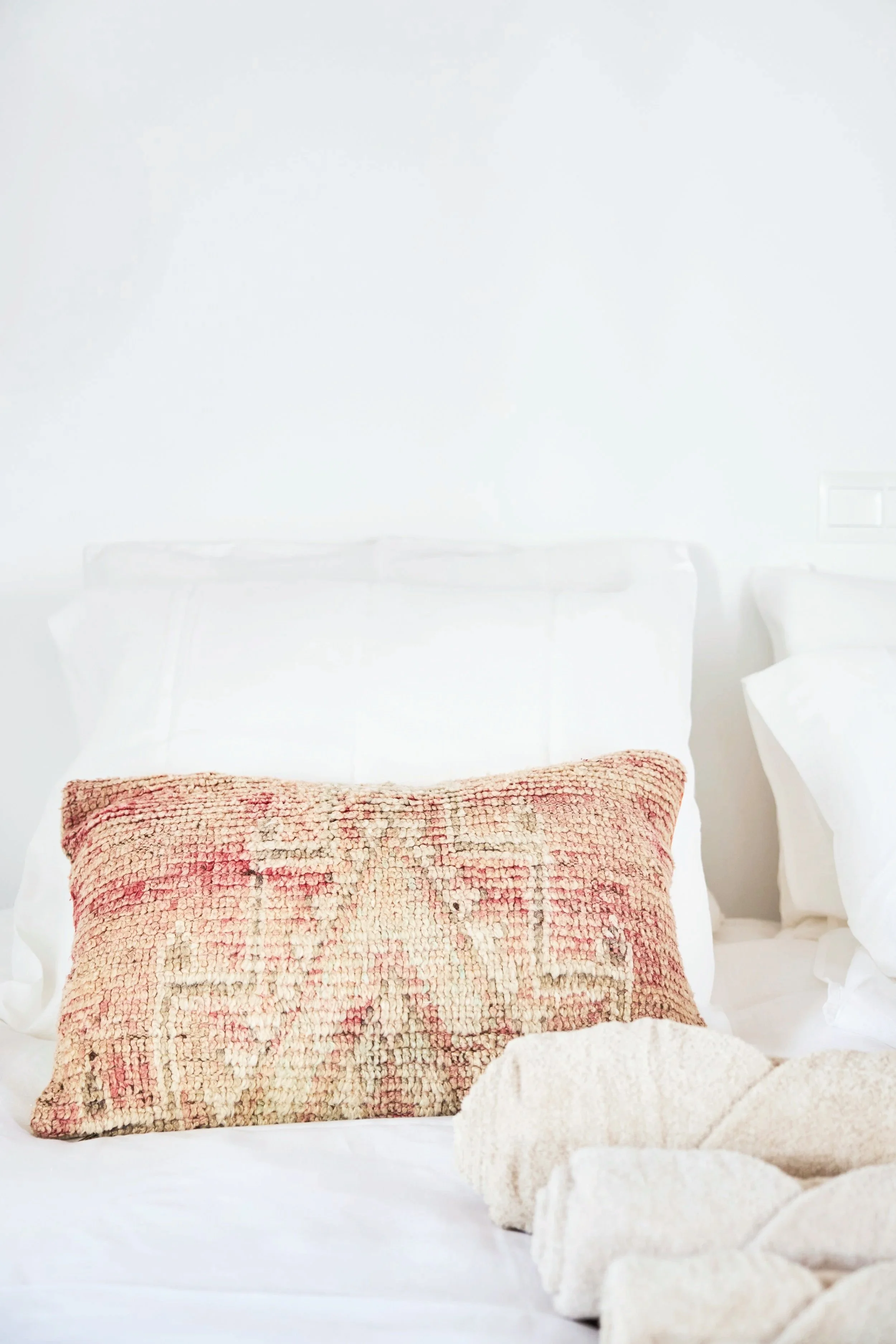 Close-up of a neatly made bed with white bedding, featuring a decorative pillow with a star design and beige textured towel or blanket at the foot of the bed, against a white wall.