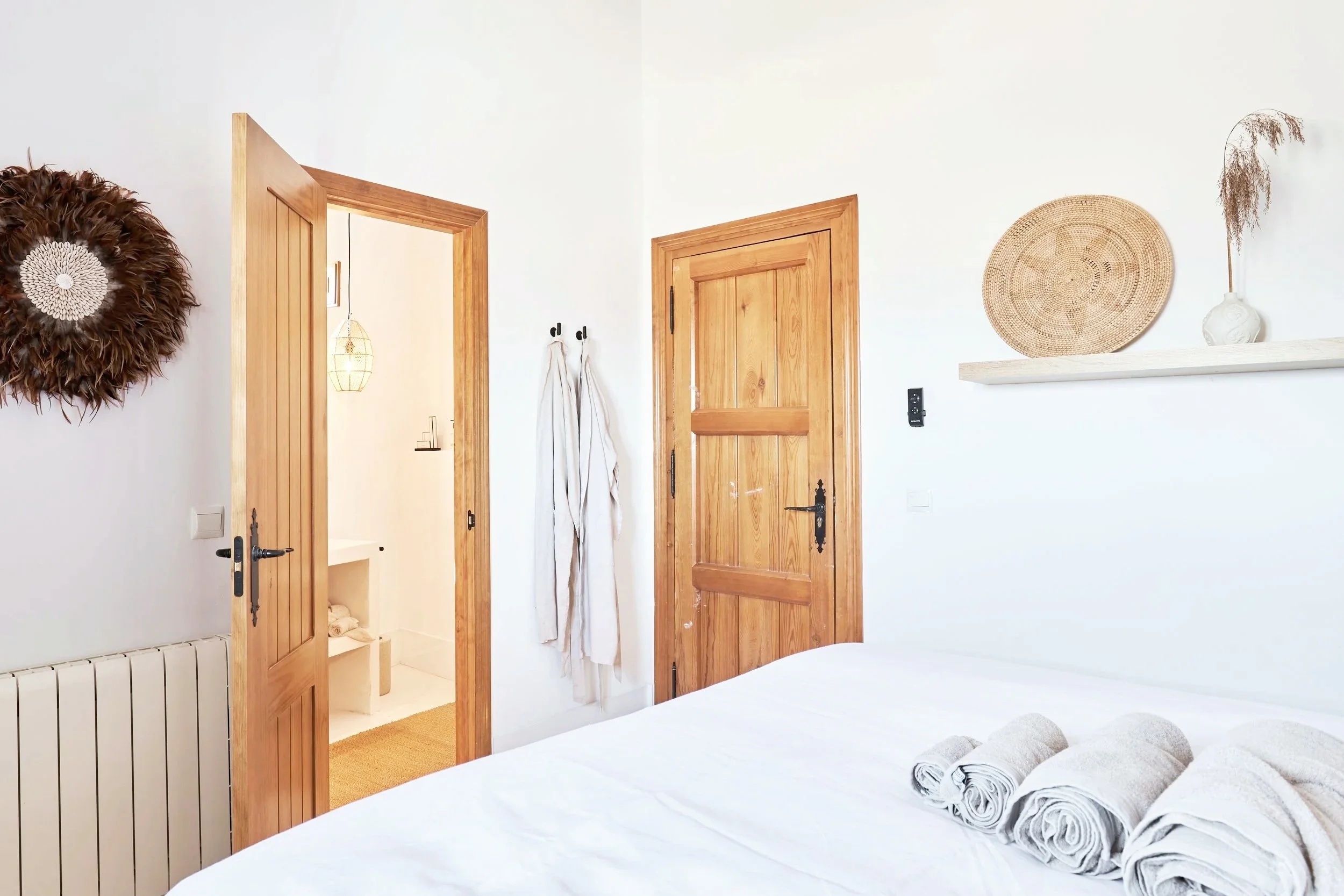 A bedroom with a white bed, three rolled towels on the bed, wooden doors, a wall-mounted shelf with a basket and a vase with dried plant, and a doorway leading to a bathroom with a hanging light