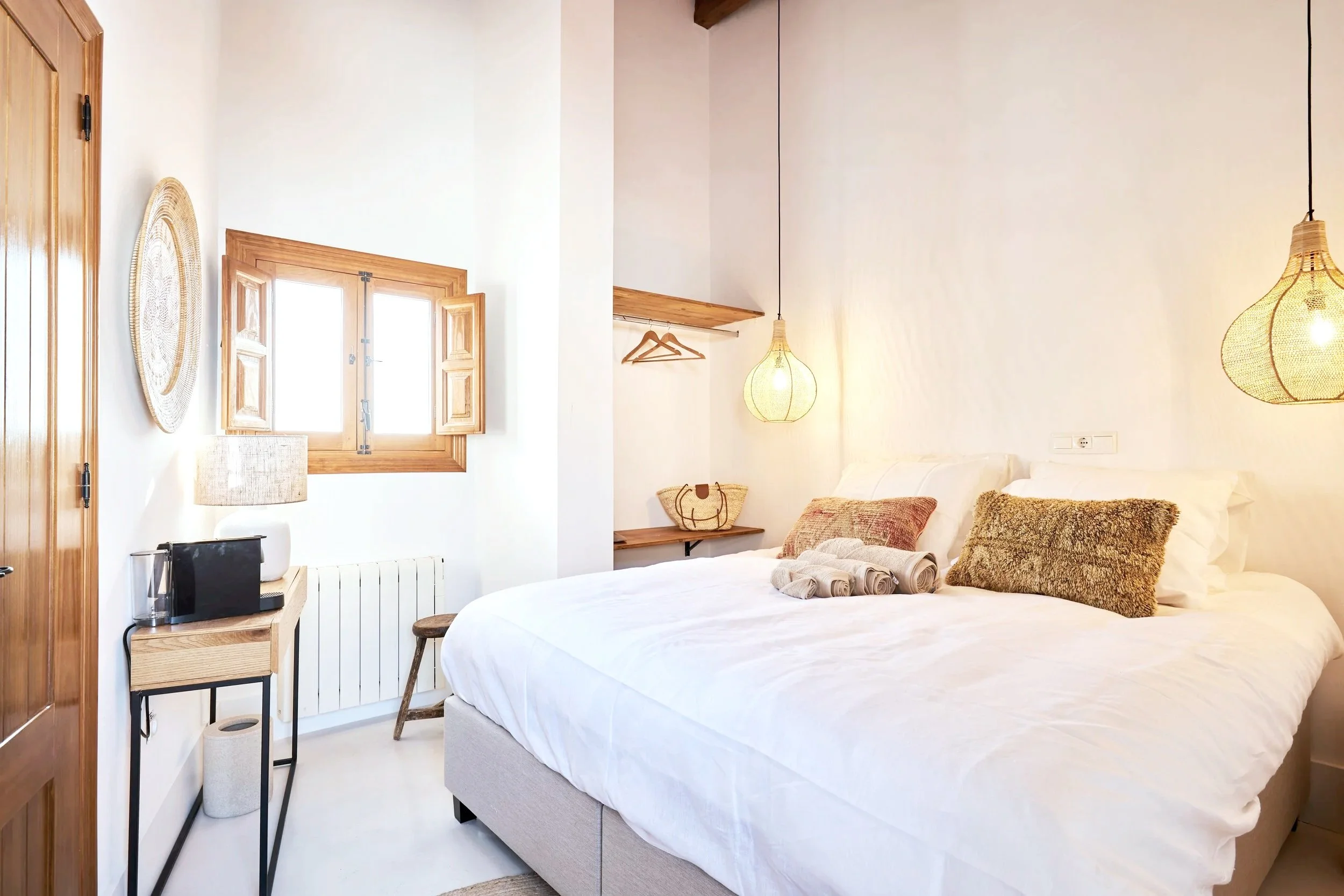 Cozy bedroom with white bed, decorative pillows, wooden window with shutters, hanging pendant lights, and minimalist furniture.