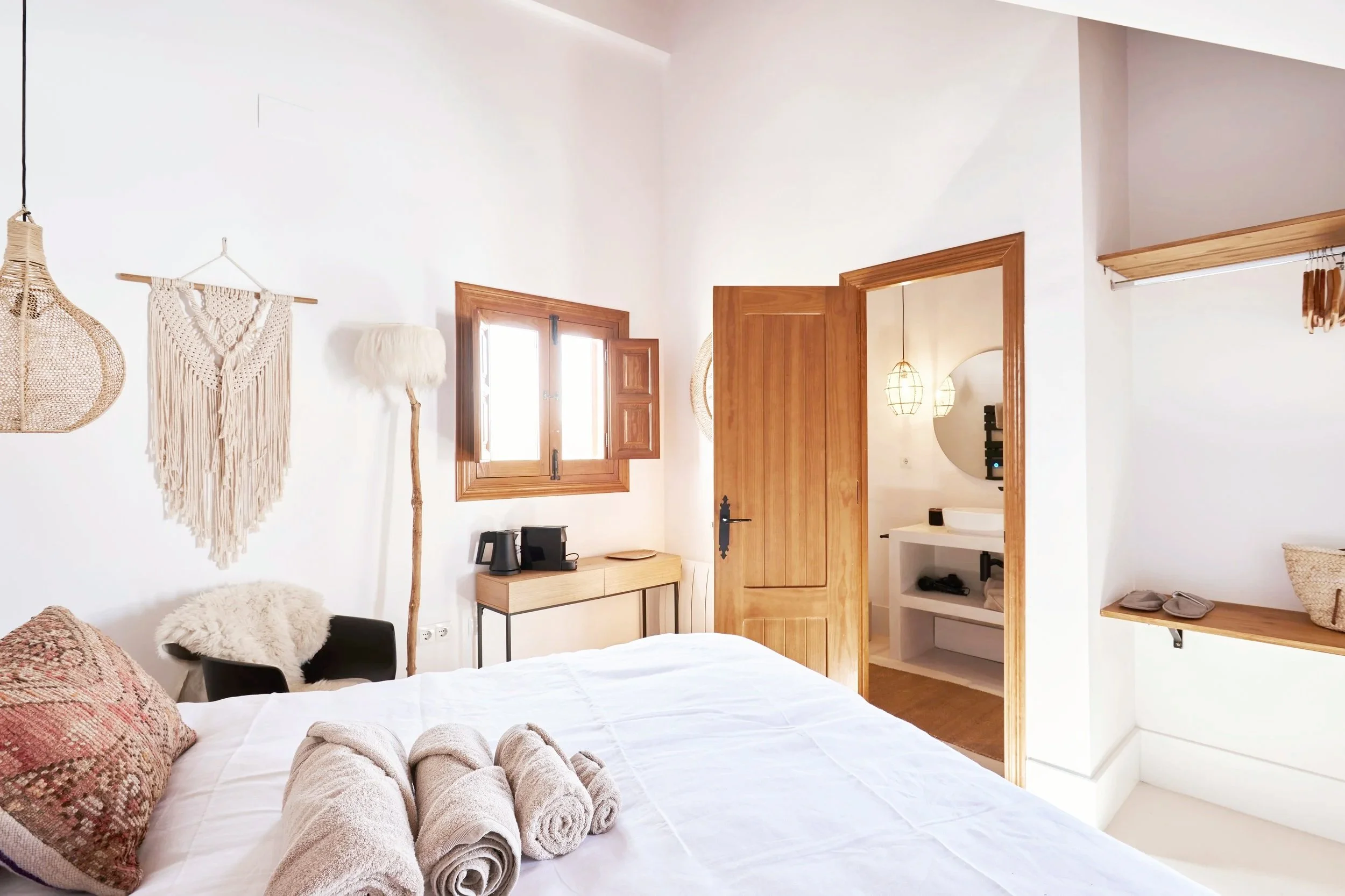 A cozy bedroom with a white bed, towels, and a decorative pillow. Wall decorations include a woven hanging, a fluffy floor lamp, and a wicker basket. Small furniture includes a black armchair with a fur throw, a wooden bench, and a door leading to an