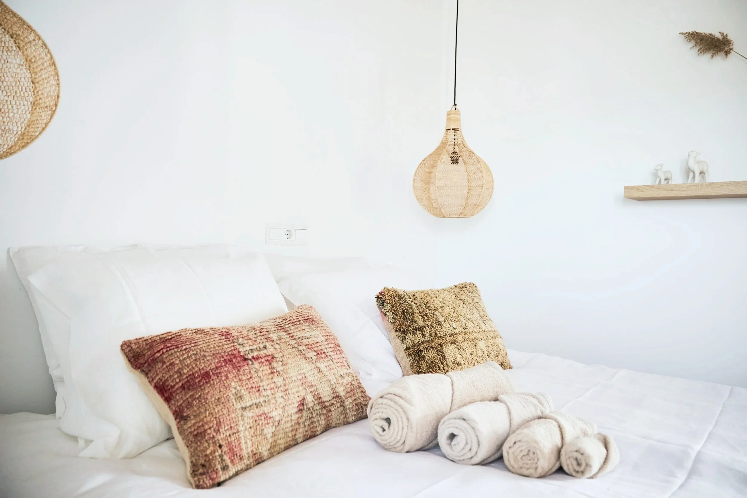 A neatly made bed with white linens and two textured accent pillows, beside rolled towels, in a bedroom with minimal decor, a hanging wicker light, and a floating wooden shelf with decorative figures.