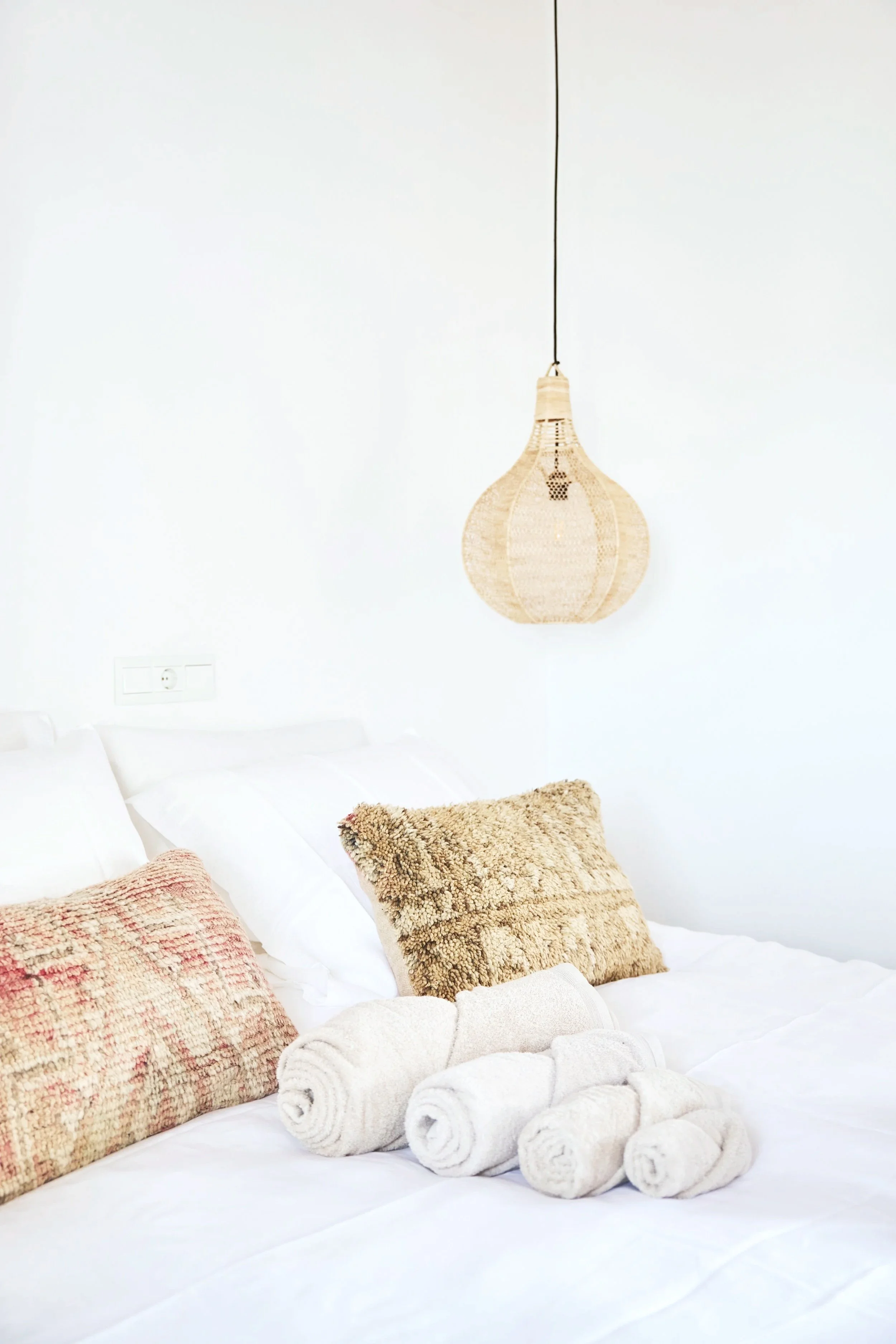 A neatly made bed with white linens, several textured pillows, and rolled towels on top. A woven pendant light hangs from the ceiling against a plain white wall.