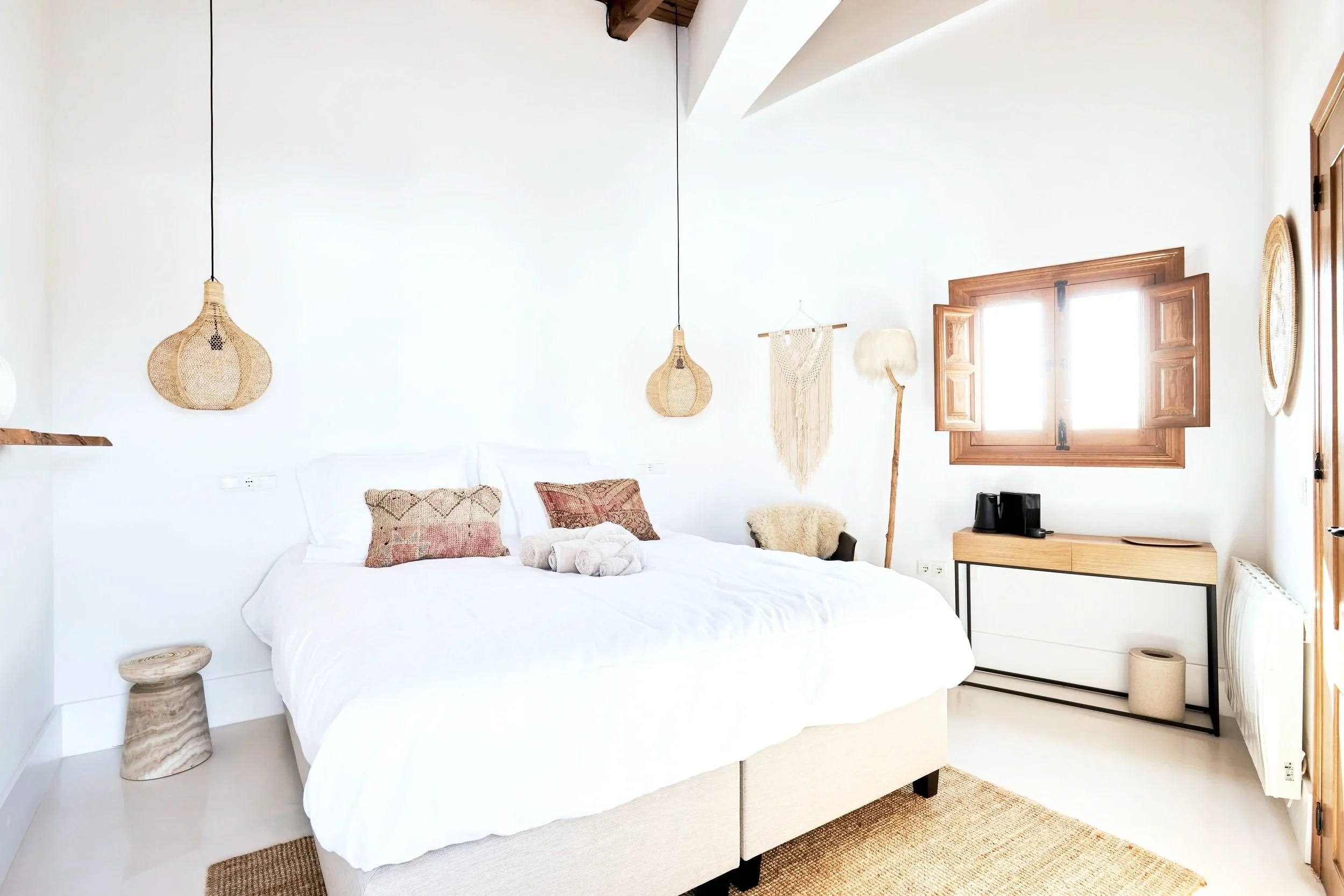A cozy bedroom with a large bed covered in white bedding, decorated with patterned throw pillows and rolled towels. There are hanging woven lamps, a macrame wall hanging, a floor lamp with a fabric shade, and wooden window shutters allowing light int