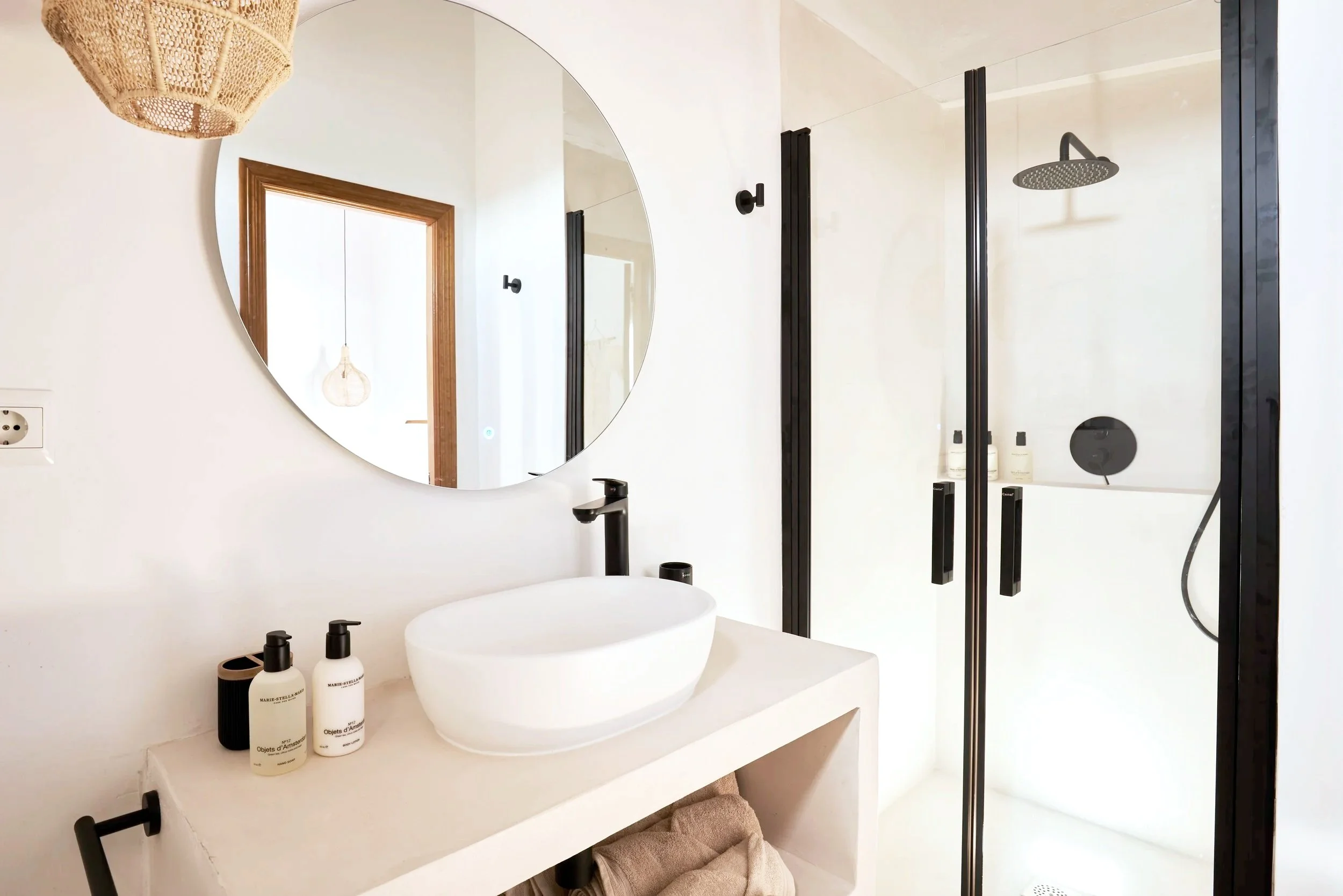 Modern bathroom with round mirror, white vessel sink, black faucet, and black fixtures. Glass shower stall with black framing and a rain showerhead.